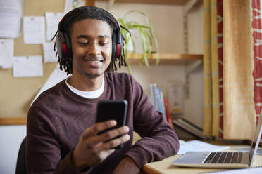 Male University Or College Student Working On Laptop Wearing Wireless Headphones Looking At Mobile Phone At Desk In Room - INGF12701