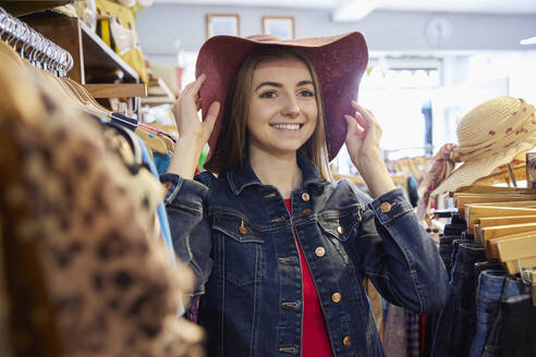 Junge Frau kauft gebrauchte nachhaltige Kleidung aus zweiter Hand Charity Shop Trying On Hat - INGF12704