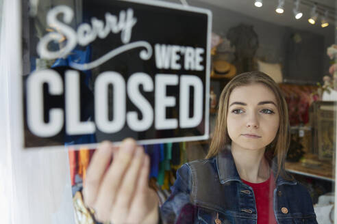 Sad Female Owner Of Small Business Turning Round Closed Sign In Shop Window - INGF12709