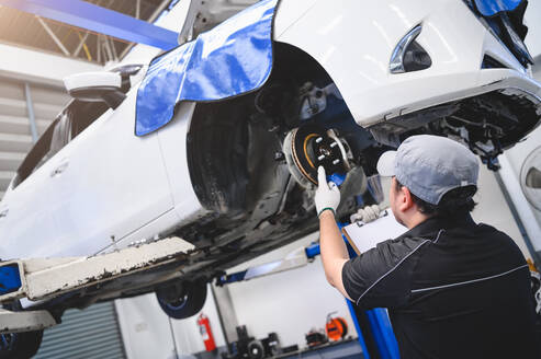 Asian male car technician car maintenance for customers according to specified vehicle maintenance checklist. Disk brake pad wear automotive repairing on vehicle. Safety inspection check service - INGF12780