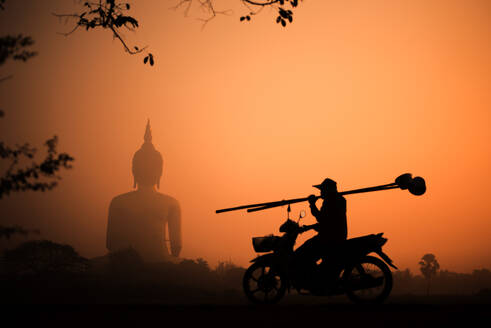 Silhouette big Buddha with the golden sky on the sunset and silhouette of man riding a motorcycle holding bamboo stick afterwork. - INGF12783