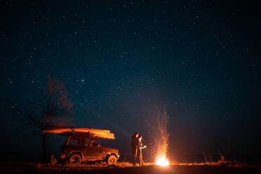Happy couple man and woman standing in front burning bonfire under starry sky, enjoying quiet night. Happy couple man and woman standing in front burning bonfire - INGF12813