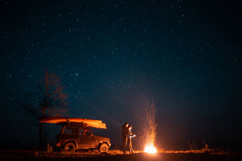 Happy couple man and woman standing in front burning bonfire under starry sky, enjoying quiet night. Happy couple man and woman standing in front burning bonfire - INGF12813