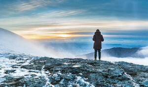 A person standing on a snow covered cliff over the breathtaking view of the mountains under the sunset. Person standing on a snow covered cliff over the breathtaking view of mountains under the sunset - INGF12819