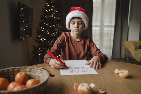 Junge mit Weihnachtsmannmütze schreibt auf Papier am Tisch - ALKF00653