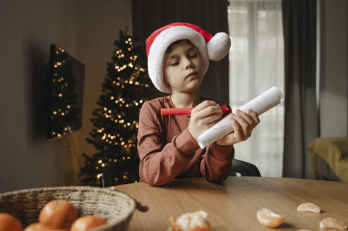 Junge mit Weihnachtsmannmütze, der am Tisch im Haus auf Papier schreibt - ALKF00654