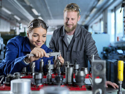 Smiling trainee learning from metal worker to check CNC tool with caliper at factory - CVF02556