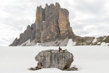 Wanderer sitzt auf einem Felsen umgeben von Schnee vor einem Berg - MMPF00921
