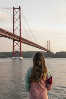 Junge Frau bewundert den Fluss Tejo mit der Brücke des 25. April bei Sonnenuntergang - MMPF00927