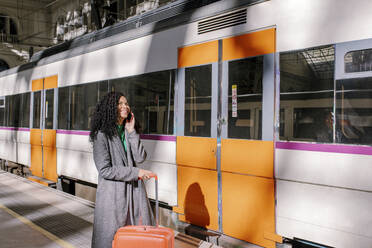 Glückliche Frau mit Koffer, die auf dem Bahnsteig mit ihrem Smartphone spricht - MMPF00931