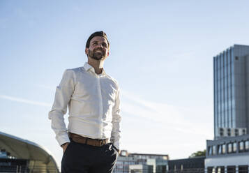 Happy businessman standing with hands in pockets on sunny day - UUF30462