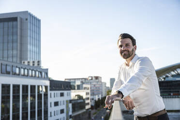 Lächelnder Geschäftsmann am Geländer stehend an einem sonnigen Tag - UUF30470