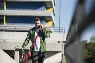 Smiling man talking on smart phone and walking with bicycle in front of building - UUF30477