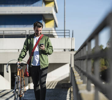 Lächelnder Mann, der ein Smartphone benutzt und mit einem Fahrrad vor einem Gebäude spazieren geht - UUF30479