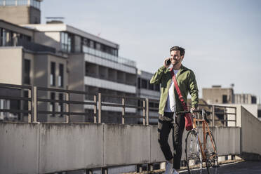 Smiling man talking on smart phone and walking with bicycle near railing - UUF30480