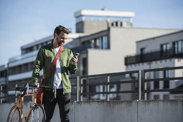 Happy man using smart phone and walking with bicycle near railing on sunny day - UUF30481