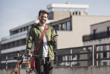 Lächelnder Mann mit Smartphone und Fahrrad an einem sonnigen Tag - UUF30482