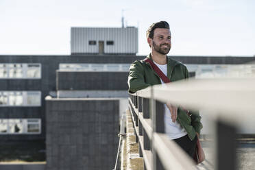 Happy man standing with bag near railing on sunny day - UUF30493