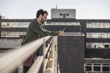 Glücklicher Mann, der sein Smartphone benutzt und sich auf ein Geländer vor einem Gebäude stützt - UUF30494