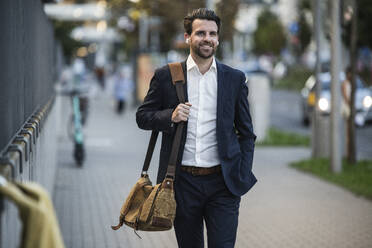 Thoughtful businessman with crossbody bag walking on footpath - UUF30514