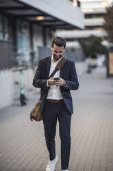 Businessman with crossbody bag using smart phone walking on footpath - UUF30515