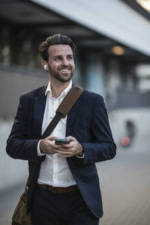 Lächelnder Geschäftsmann mit Mobiltelefon auf dem Fußweg - UUF30517