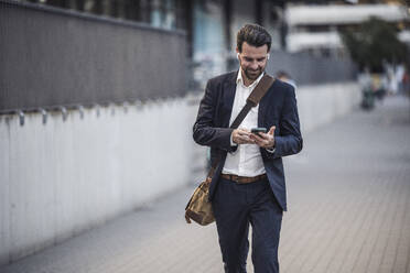 Businessman using smart phone walking on footpath - UUF30518