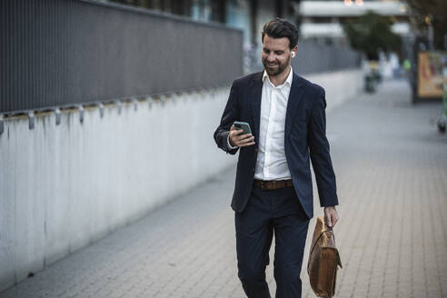 Businessman with briefcase using mobile phone walking on footpath - UUF30519