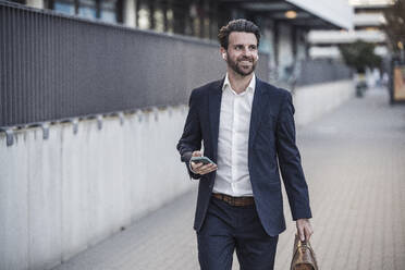 Lächelnder Geschäftsmann mit Smartphone auf dem Gehweg - UUF30520