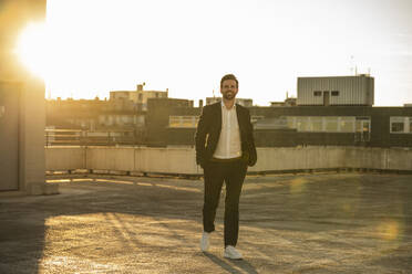 Businessman in blazer walking on terrace at sunset - UUF30525
