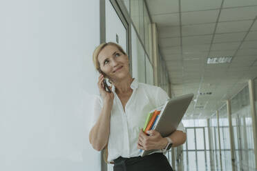 Lächelnde Geschäftsfrau, die einen Laptop hält und mit einem Smartphone im Büro telefoniert - OSF02162