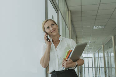 Smiling businesswoman holding laptop and talking on smart phone in office - OSF02162