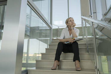 Kontemplative Geschäftsfrau auf den Stufen sitzend mit Laptop und Büchern im Büro - OSF02167