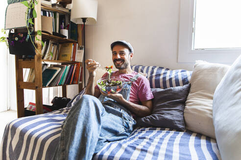 Lächelnder Mann entspannt sich und isst Salat auf dem Sofa zu Hause - PCLF00706