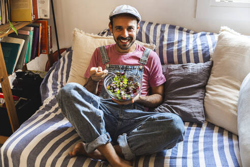 Glücklicher Mann isst Salat auf dem Sofa zu Hause - PCLF00707