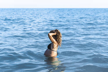 Schwangere Frau, die ihre Haare berührt und im Meerwasser steht - PCLF00709