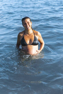 Glückliche schwangere Frau mit Händen auf dem Bauch im Wasser stehend - PCLF00712