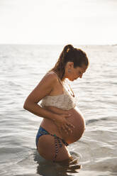 Pregnant woman with hands on stomach standing in water - PCLF00717