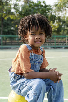 Nachdenklicher Junge sitzt auf einem Basketball auf dem Sportplatz - IKF01239