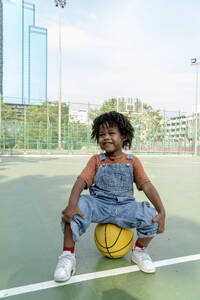 Verspielter Junge sitzt auf einem Basketball auf dem Sportplatz - IKF01243