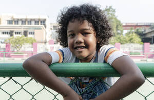 Fröhlicher Junge lehnt am Zaun eines Sportplatzes - IKF01246