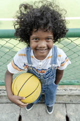 Lächelnder Junge hält Basketball in der Nähe des Zauns am Sportplatz - IKF01247