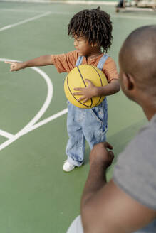 Vater mit Sohn hält Basketball und zeigt auf Sportplatz - IKF01253