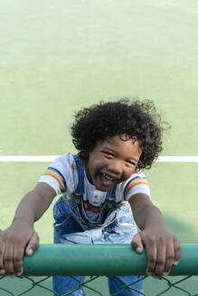 Lächelnder Junge hält Zaun am Sportplatz - IKF01262