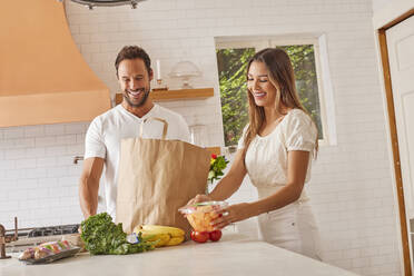 Smiling couple with paper shopping bag and groceries in kitchen - TETF02227