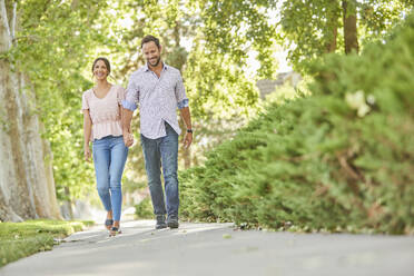 Smiling couple holding hands, walking on treelined sidewalk - TETF02239