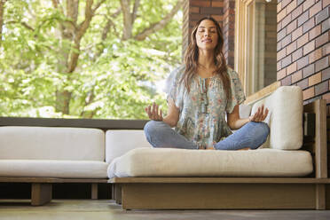 Mid adult woman meditating on sofa on patio - TETF02245