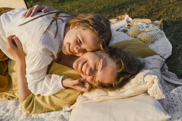 Glückliche Mutter, die ihre Tochter auf einer Picknickdecke bei Sonnenuntergang umarmt - VBUF00405