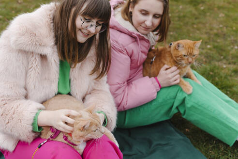 Zwei junge Frauen halten Katzen und sitzen im Gras im Park - EHAF00107