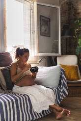 Pregnant woman eating oatmeal on sofa at home - PCLF00726